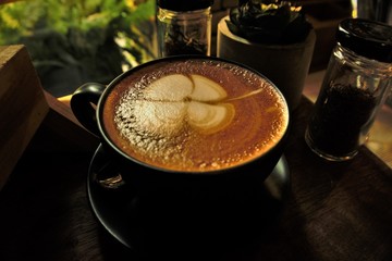 Close up black coffee cup with latte art foam as a clover leaf on wooden table and blurred background, Hua Hin Thailand.