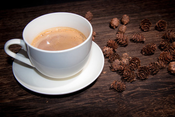 white cup of coffee on wood table and some pine cones on the background.