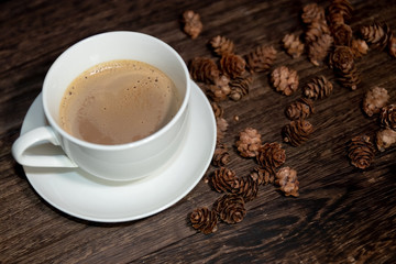 white cup of coffee on wood table and some pine cones on the background.