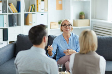 Mature blonde female psychologist sitting in front of young troubled couple and consulting them in office - obrazy, fototapety, plakaty