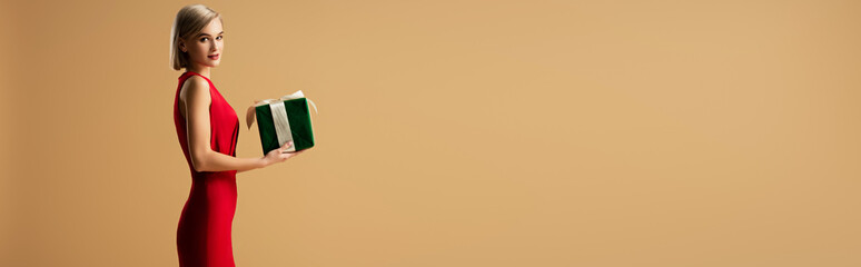 panoramic shot of beautiful young woman in red dress holding gift box isolated on beige