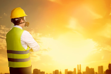 Rear view of asian worker man in safety vest, gloves, yellow helmet and protective mask standing and looking at city