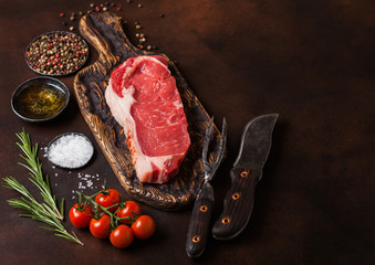 Raw sirloin beef steak on old vintage chopping board with knife and fork on rusty background. Salt and pepper with fresh rosemary and tomatoes.Space for text