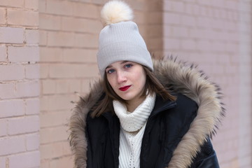 Exquisite blue-eyed young woman in fur trimmed winter coat and pom pom hat standing in front of pink brick wall