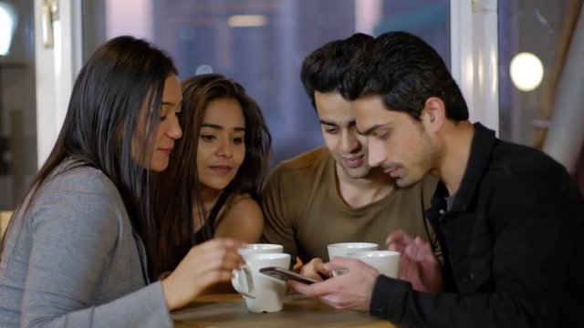 Indian young friends take photos selfie on a phone as they pose and pout and then review and criticize the photo over a coffee together on a cafe talking chatting and talking 