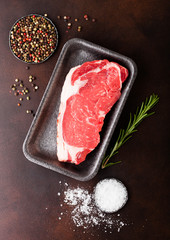 Raw sirloin beef steak in plastic tray with salt and pepper and fresh rosemary on rusty background.
