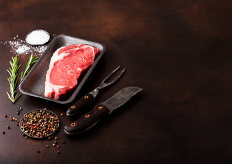 Raw sirloin beef steak in plastic tray with knife and fork on rusty background. Salt and pepper with fresh rosemary