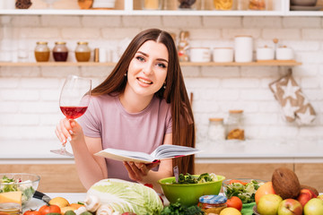 Evening at home. Cooking hobby. Smiling young woman with red wine glass. Book of healthy recipes in hands.