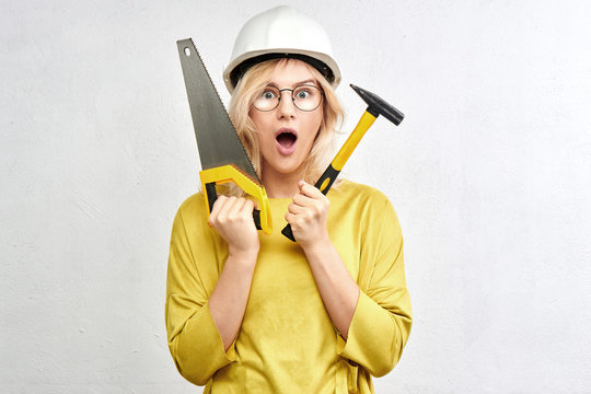 Surprised hot builder engineer with open mouth, tools, construction helmet, glasses, saw and hammer in white studio. Concept repair and service