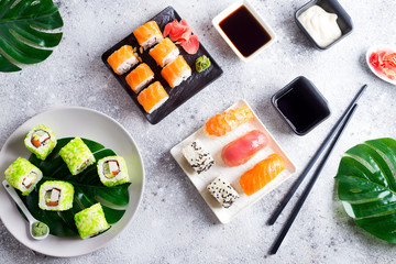 Various Fresh and delicious sushi set on black and white slate with metal and slate sticks, sauce and green leaves on light stone background , flat lay