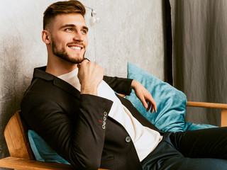Portrait of handsome smiling hipster  businessman model wearing casual black suit. Fashion stylish man posing sitting in chair in luxury interior. Thinking