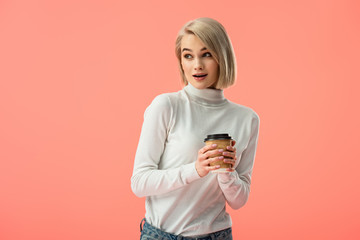 Fototapeta premium shocked blonde girl holding paper cup with drink isolated on pink
