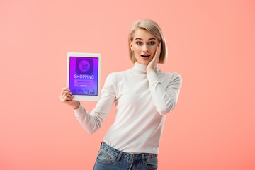 Naklejka premium surprised blonde woman holding digital tablet with shopping app on screen isolated on pink