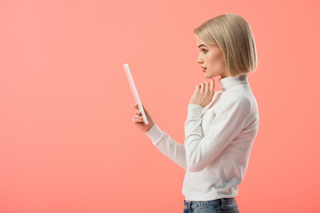 surprised blonde woman holding digital tablet isolated on pink