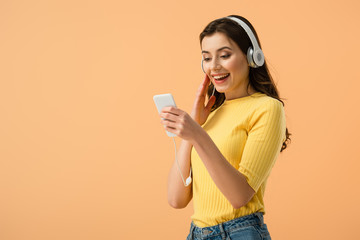 Joyful brunette girl in headphones using smartphone with smile isolated on orange