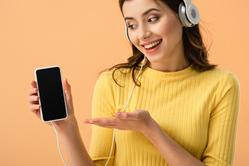 Happy brunette girl in headphones showing smartphone with blank screen isolated on orange