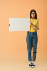 Fototapeta na wymiar Pensive brunette girl in jeans and jumper holding blank placard on orange background