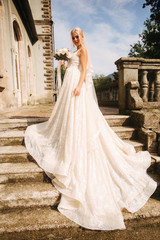 Obraz premium Gorgeous bride in wedding dress standing on the stairs by the palace. She hold a bouquet of flowers