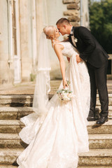 Groom and bride stand on stairs. Fashion model in elegant clothes. White long dress and stylish suit