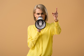 Serious blonde woman in yellow shirt talking in megaphone and pointing with finger isolated on beige