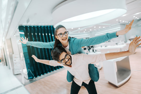 Cheerful multicultural couple with eyeglasses dressed casual having piggyback at tech store. - Powered by Adobe