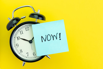 Sticky post with handwriting the word Now stick on alarm clock on solid yellow background with copy...
