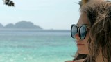 Close-up slow motion portrait of beautiful european young woman in sunglasses, hair blowing in wind on tropical beach, blue sea on the background. Travel concept.