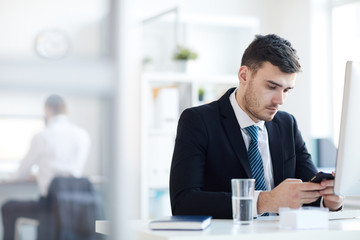 Serious young businessman sitting by workplace in front of computer and texting in smartphone