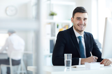 Young successful businessman in formalwear sitting by desk in front of computer monitor and organizing work
