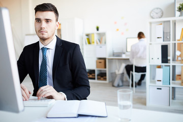 Young serious broker in elegant suit looking at computer screen while analyzing online data by workplace