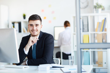 Cheerful and successful young broker in suit sitting by workplace in front of computer during online work