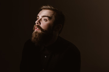 Fototapeta premium Sepia portrait of a pensive man with a beard and stylish hairdo dressed in the black shirt on the dark background