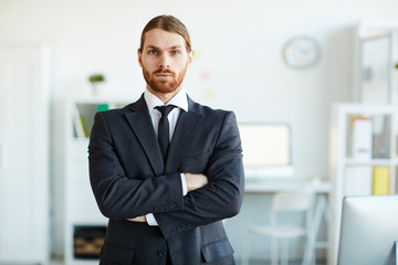 Young bearded business employee in elegant suit crossing arms on chest while standing in front of camera