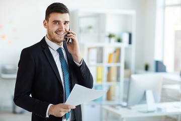 Happy young successful banker in formalwear talking to one of clients on the phone in office