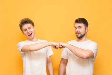 Studio shot of o bearded man and teenager, give fist bump, agree to work together, wear a white T-shirt, look at the camera and smile on the yellow background.
