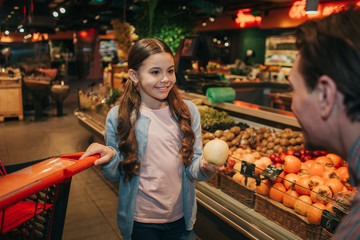 Young parent and daughter in grocery store. She hold apple in hand and hand of trolley. Girl look at father and smile. Man smile.