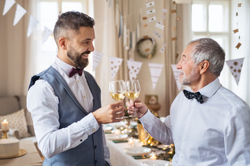 A senior and mature man standing indoors in a room set for a party, clinking glasses.