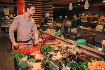 Young man in grocery store alone. He hold hand trolley and look at baskets with fruit and vegetables. Careful serious man pick best.