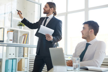 Young elegant broker with document pointing at chart on whiteboard while analyzing it at meeting