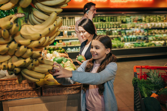 Young parents and daughter in grocery store. Girl touch yellow bananas and smile. Woman hold mango in hands. Father stand behind and talk on phone. - Powered by Adobe
