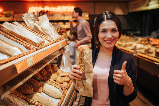 Young parents and daughter in grocery store. Positive cheerful woman hold bread in hands and show big thumb up. Fathr and daughter stand behind and pick up rolls.
