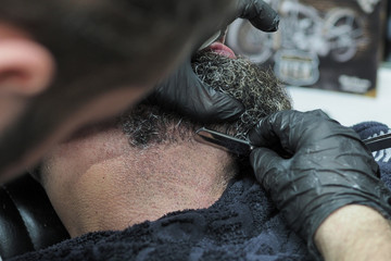Barber shaves the beard of an elderly man with gray hair sharp razor