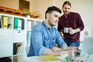 Obraz premium Two young casual employees looking at laptop display while browsing in the net and searching for new ideas