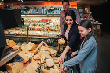 Young parents and daughter in grocery store. Mother help daughter to pick up cheese. Father stand...
