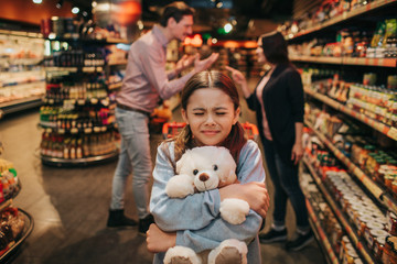 Young parents and daughter in grocery store. She sit in trolley and embrace toy bear. Girl keep eyes closed. Parents hav quarrel behind.