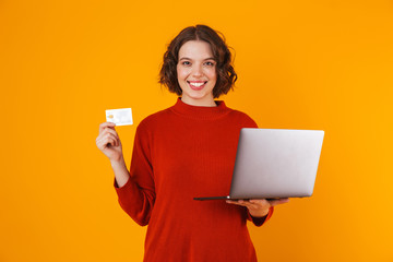 Image of brunette woman using silver laptop and credit card while standing isolated over yellow background