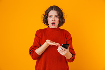 Portrait of resentful woman holding and using cell phone while standing isolated over yellow background