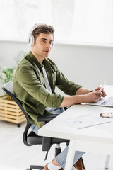 handsome businessman in headphones sitting at table with laptop, holding smartphone and listening music in office