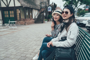Fototapeta na wymiar Attractive young travel people talking sitting on bench chair on street in solvang santa barbara us. happy two asian female tourist relaxing holding cellphone point finger to fresh new things in city