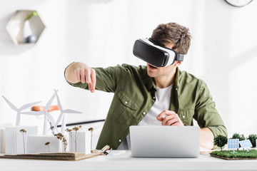 architect in black virtual reality headset sitting at table with laptop and windmills, buildings,...